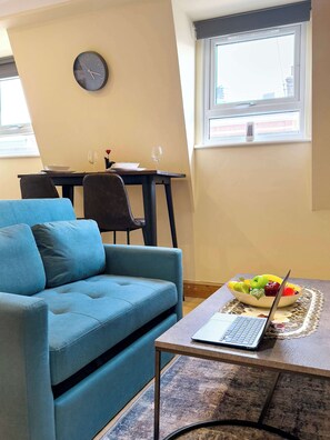 A living room with a blue sofa, a dining table with chairs, a laptop on a table, and a bowl of fruit.