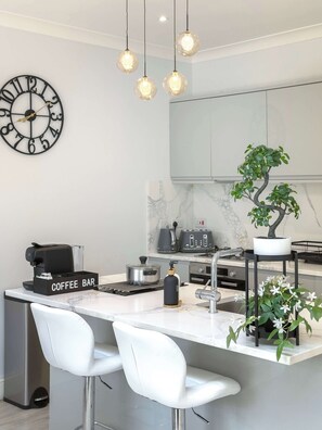 Une cuisine moderne avec un coin café, des tabourets de bar, une horloge, des luminaires suspendus et une plante en pot.