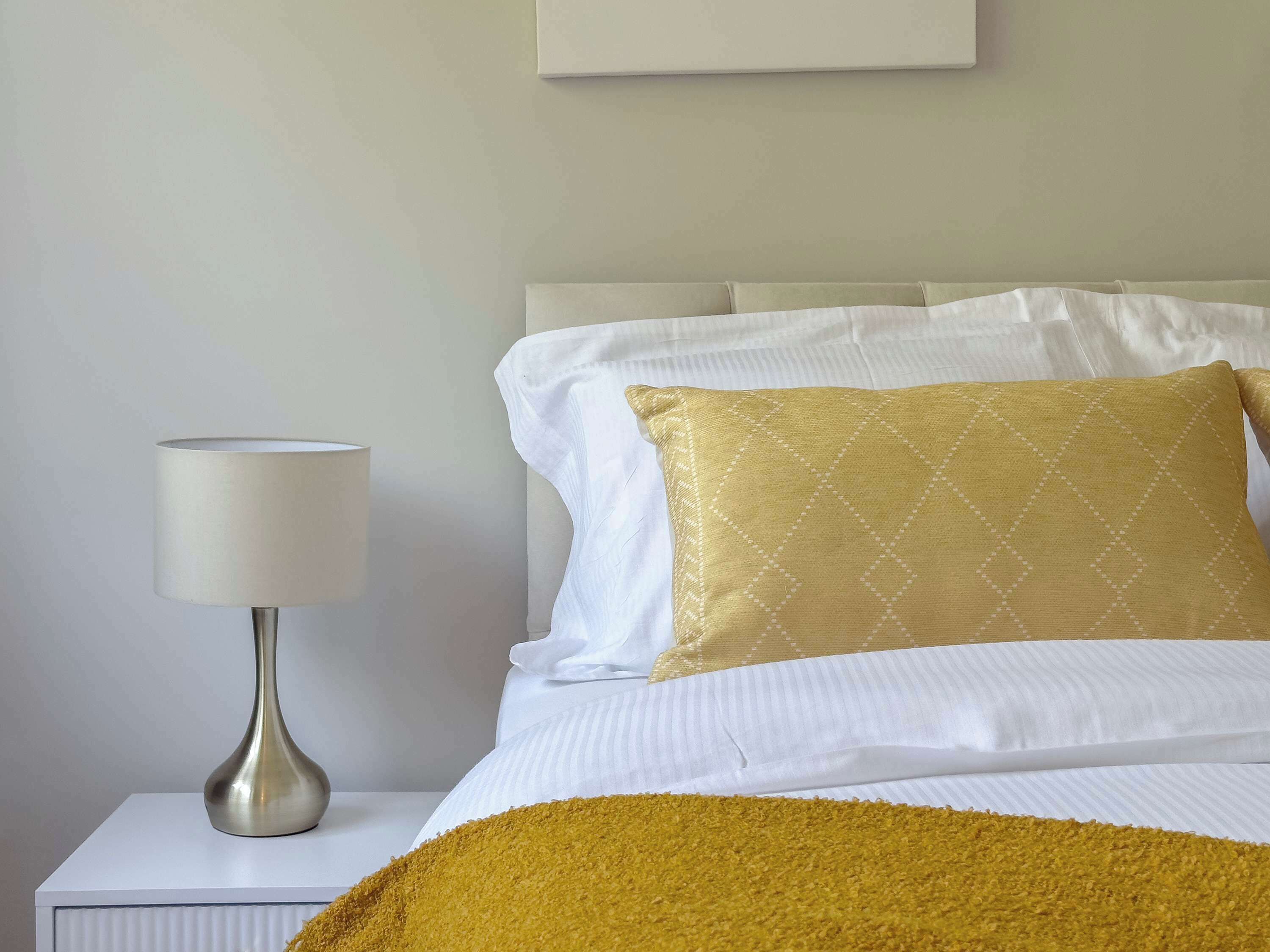 A neatly made bed with white linens and a single gold-patterned pillow, a white bedside table with a lamp, and a plain wall.
