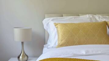 A neatly made bed with white linens and a single gold-patterned pillow, a white bedside table with a lamp, and a plain wall.