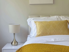 Una cama bien hecha con sábanas blancas y una única almohada con estampado dorado, una mesita de noche blanca con una lámpara y una pared lisa.