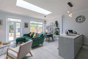 Una cocina moderna con una isla central, un comedor con sofá y sillas verdes, y un amplio ventanal que proporciona luz natural.
