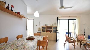 A dining area with a table covered in a patterned tablecloth, wooden chairs, a ceiling fan, and a cabinet with glass doors. - Casa Vacanze Il Bouganville (Chia)