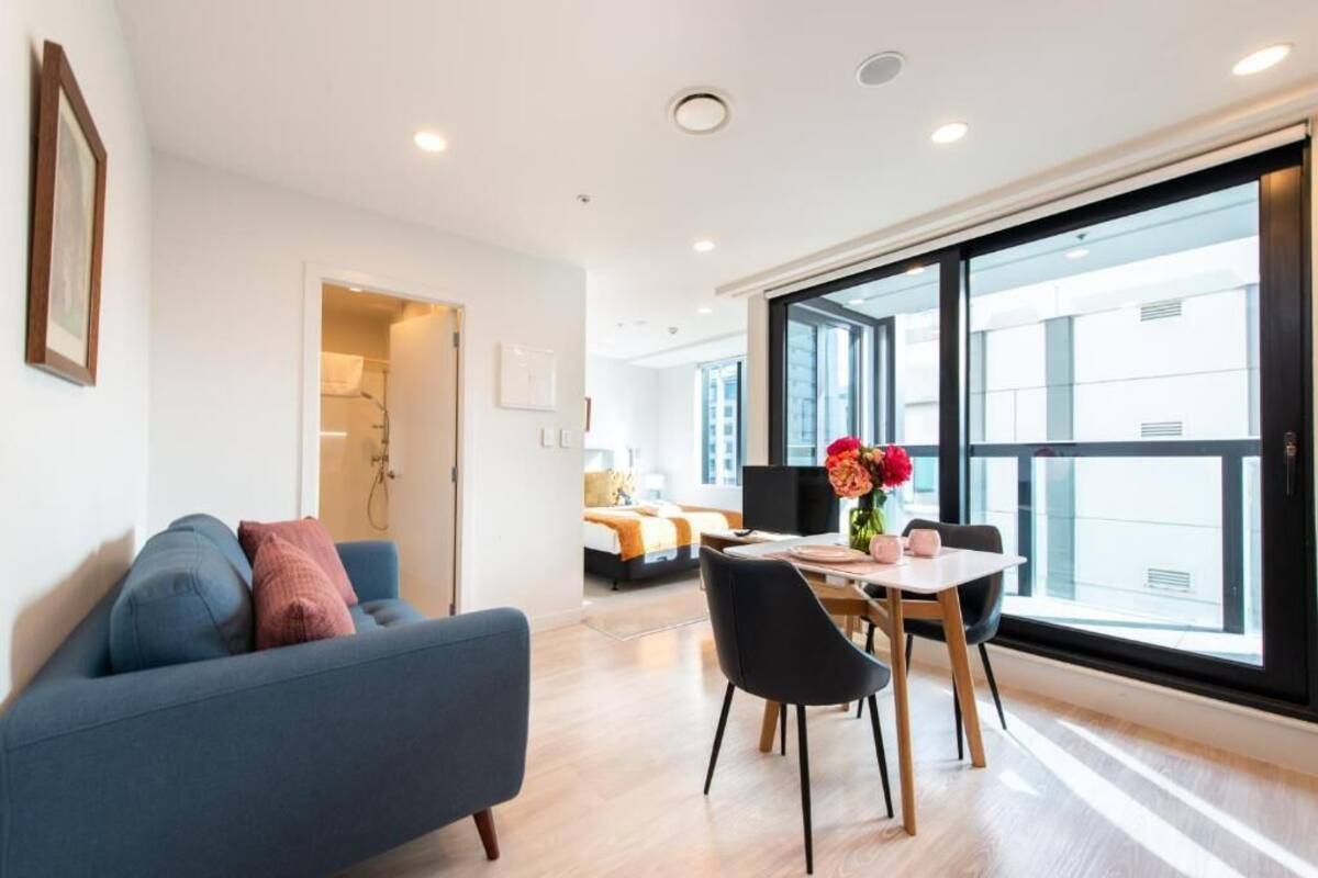 A modern living room with a sofa, dining table, and chairs. There is a large window providing natural light and a view of the city.