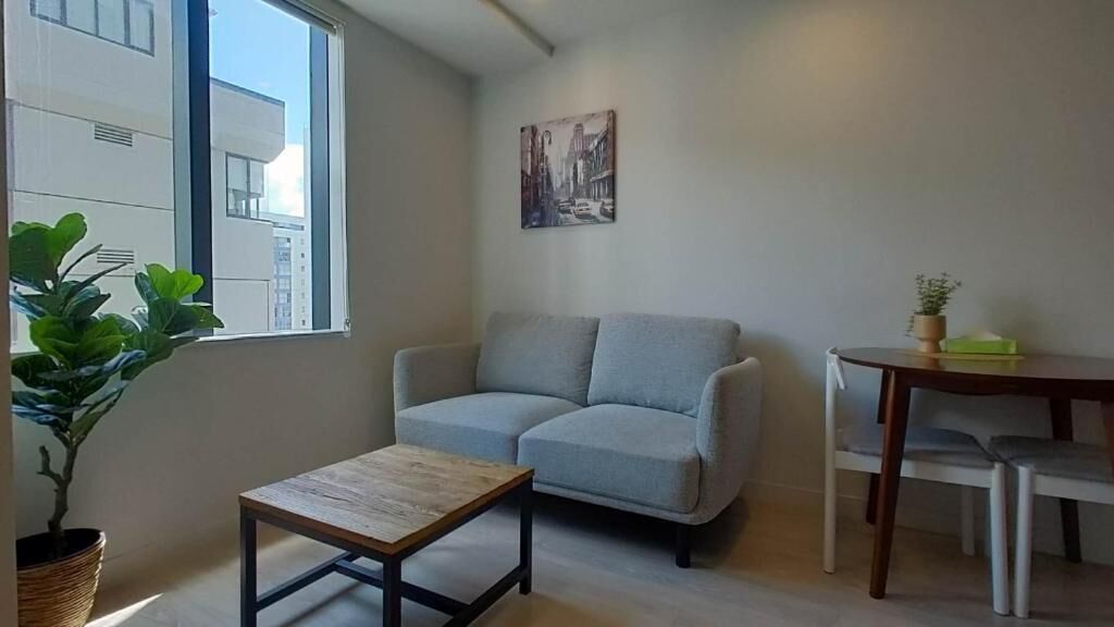 A living room with a grey sofa, a small wooden table, a dining table with chairs, a potted plant, and a cityscape painting on the wall.