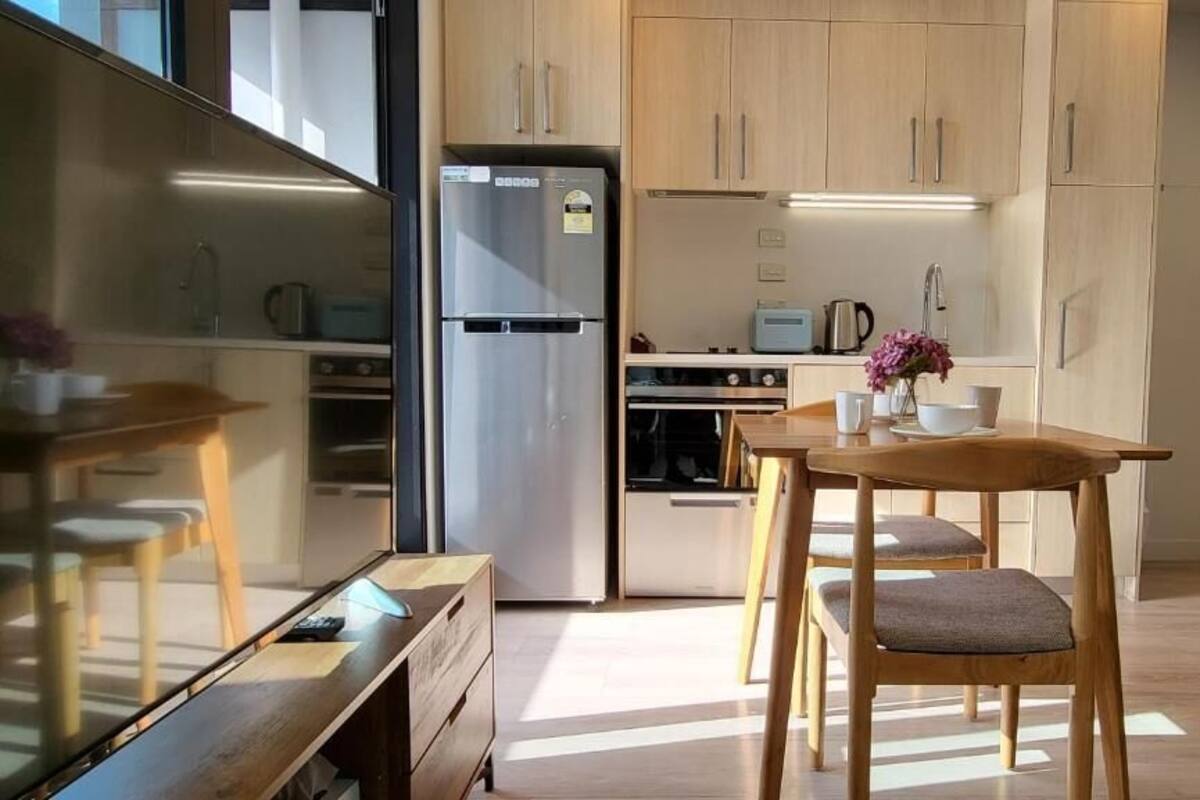 A modern kitchen with wooden cabinets, a stainless steel refrigerator, a dining table with chairs, and a small island with a sink.