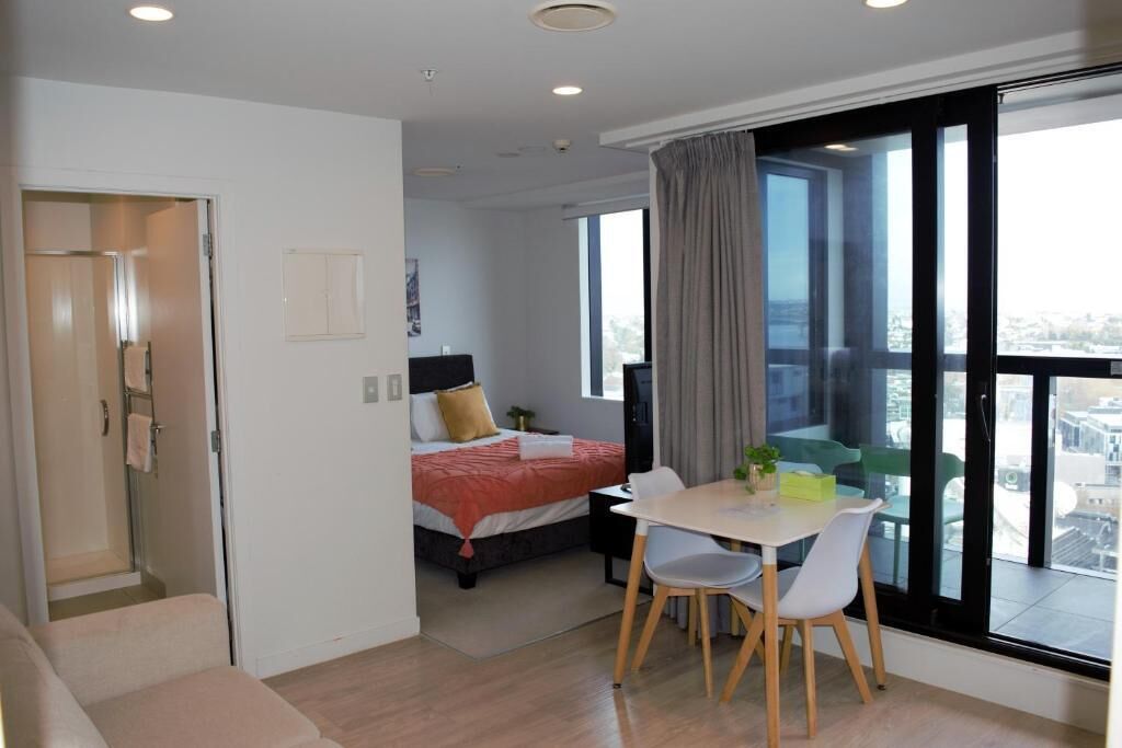 A modern hotel room with a bed, a dining area with a table and chairs, a large window with a view, and a balcony.