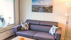 A modern living room with a grey sofa, a wooden coffee table with a yellow bowl, and a painting of a coastal landscape on the wall. - Lovely apartment for 3 people with WIFI, TV, balcony and pets allowed (Eckernförde)
