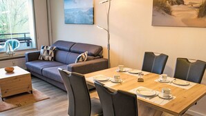A modern living room with a wooden dining table set for four, a grey sofa, and a view of the outdoors through a large window. - Lovely apartment for 3 people with WIFI, TV, balcony and pets allowed (Eckernförde)