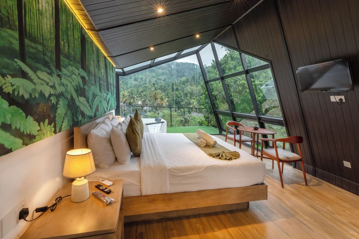 Un dormitorio moderno con una cama grande, una mesita de noche con una lámpara, un control remoto y un teléfono. Se ve una ventana con vistas a un bosque y un balcón con sillas.