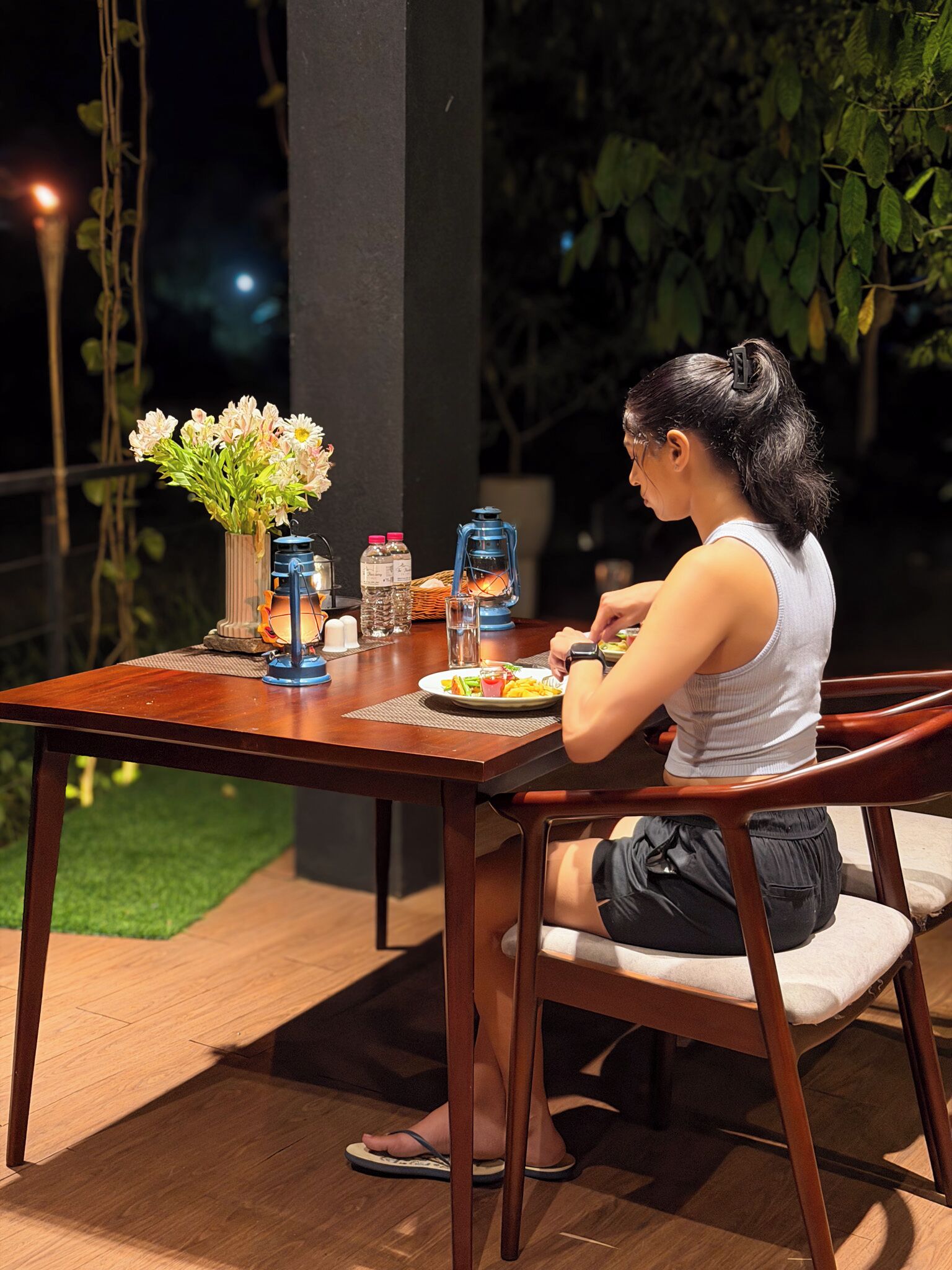 A person dining at an outdoor table with a meal, a vase of flowers, and a lantern.