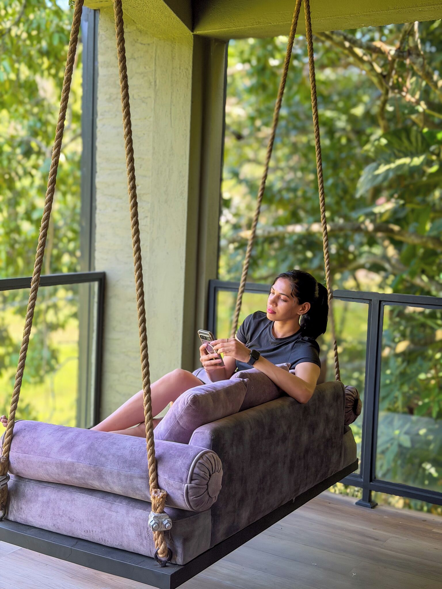A person sitting on a swing chair with ropes, in a room with a view of greenery outside.