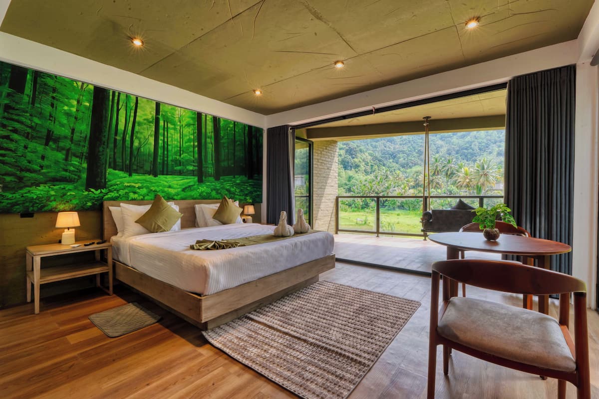 Un dormitorio con una cama grande, una mesita de noche, una silla, una mesa y vistas a un bosque a través de amplios ventanales.