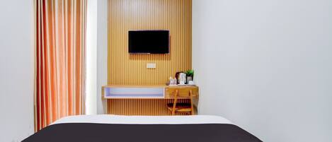 A hotel room with a bed, a mounted TV, a wooden desk with a chair, and a window with curtains.