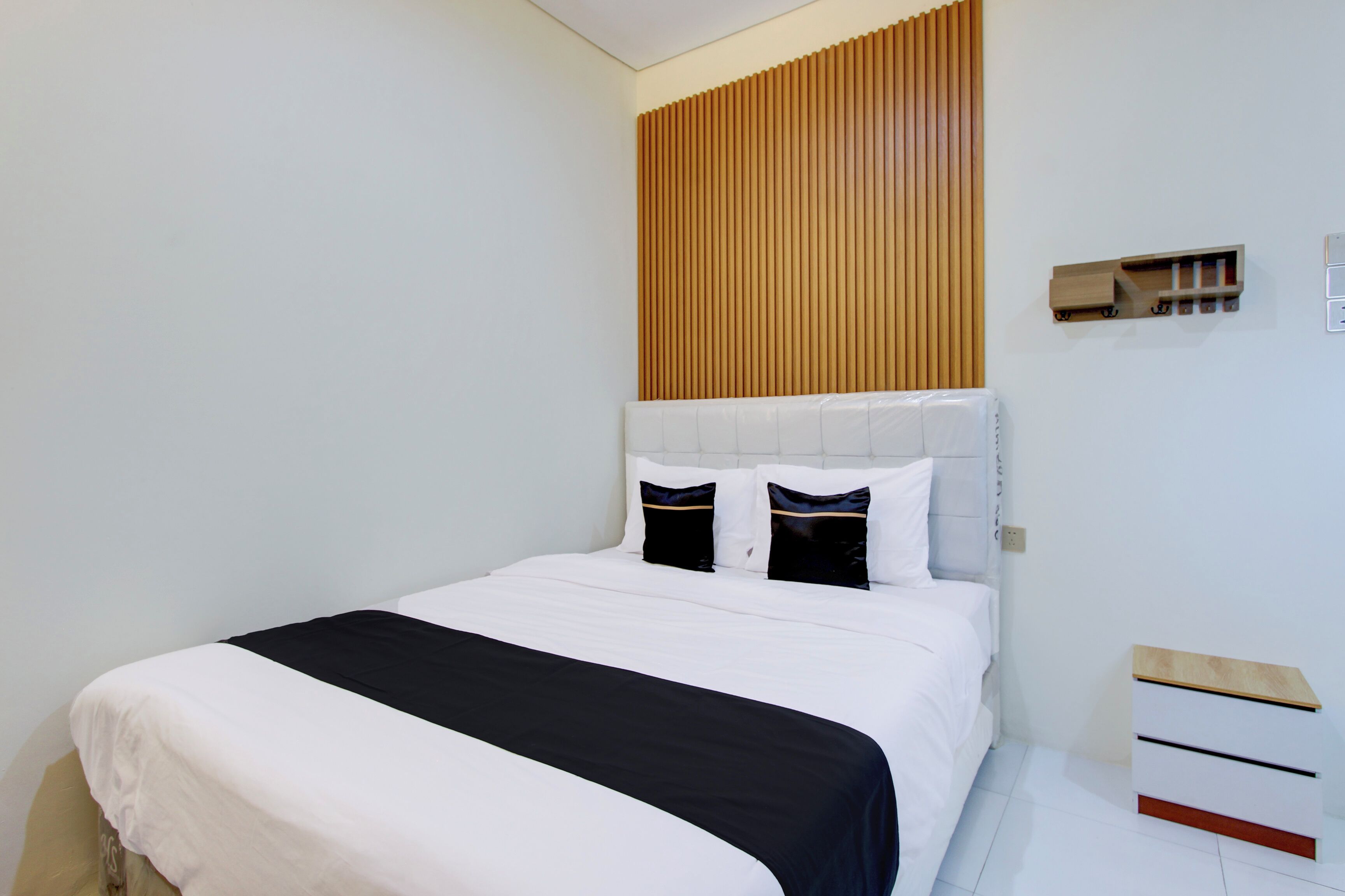 A modern hotel room with a large bed, a wooden slat headboard, a bedside table with drawers, and a small shelf with items.