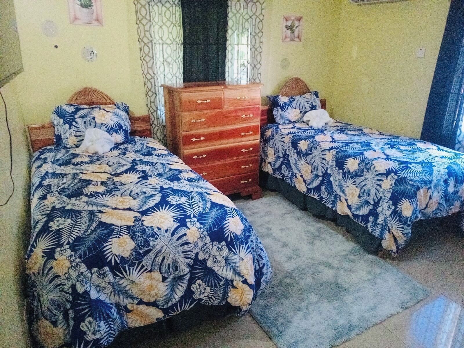 A bedroom with two beds, each with a floral bedspread, a wooden dresser, and a small potted plant on the dresser.
