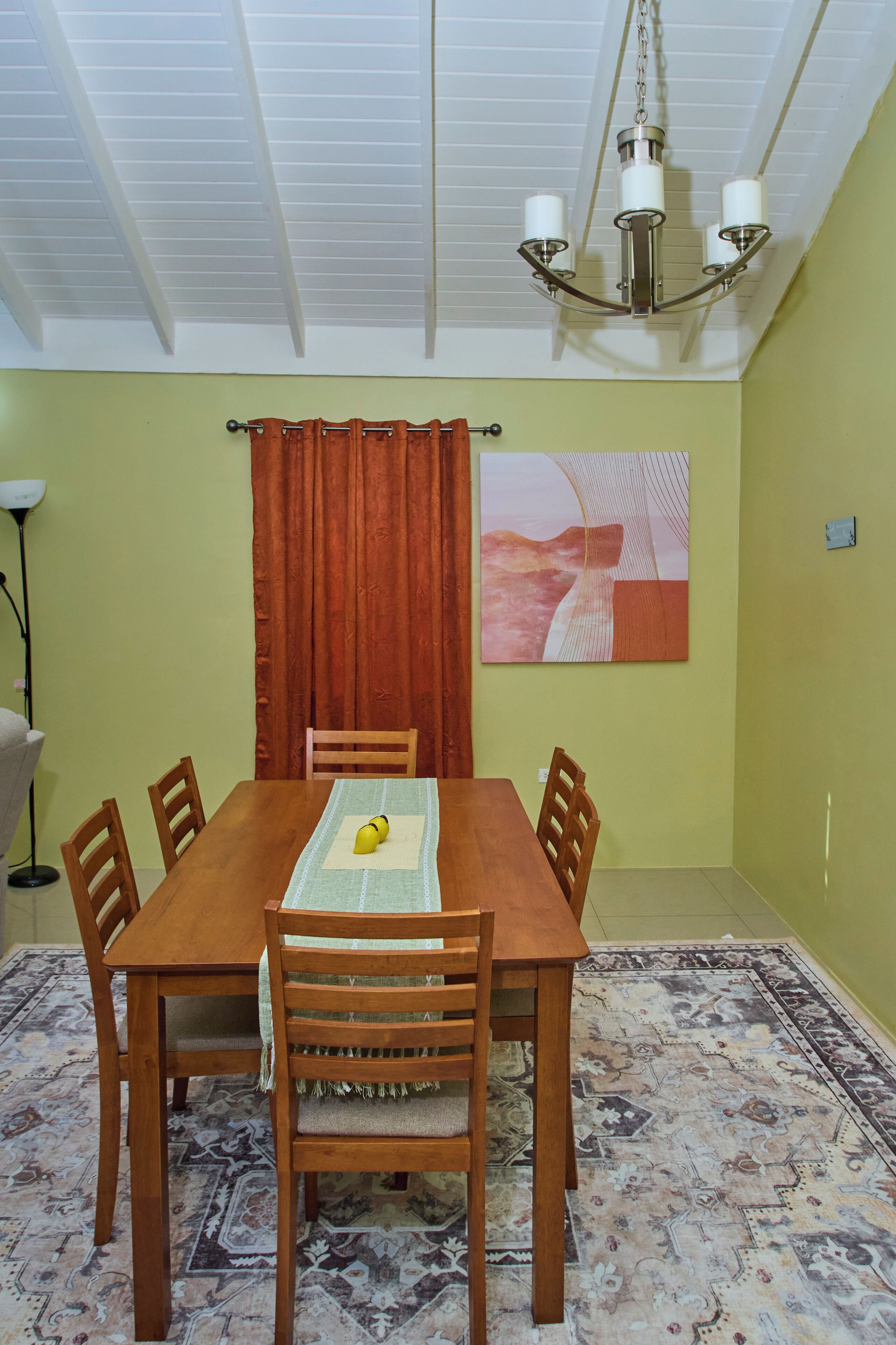 A dining area with a wooden table, six chairs, a chandelier, a patterned rug, a red curtain, and a wall art piece.