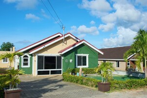 Una casa de dos pisos con fachada verde, techo rojo y entrada pavimentada.