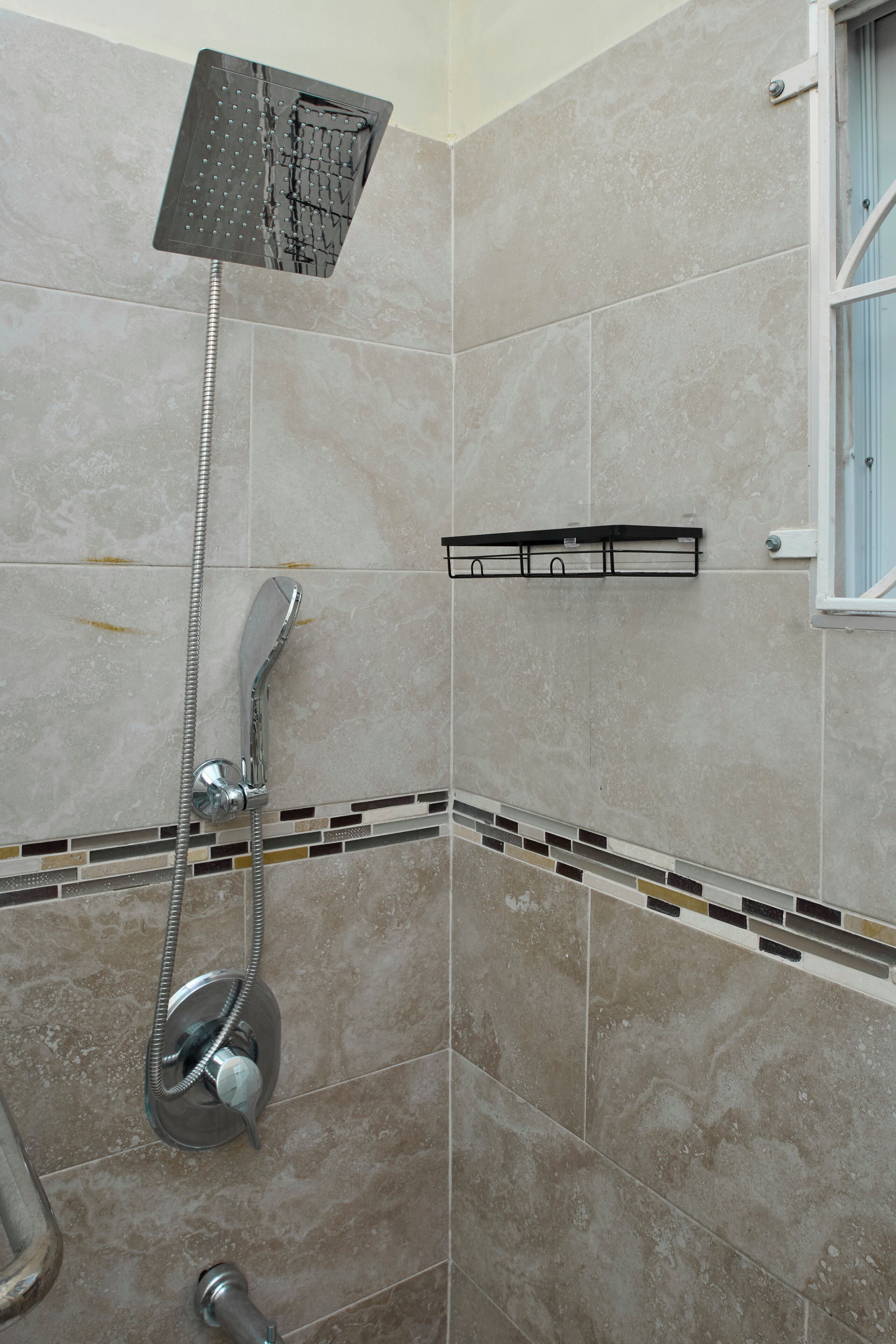 A shower with a handheld showerhead and a fixed overhead showerhead, tiled walls, and a small shelf with toiletries.