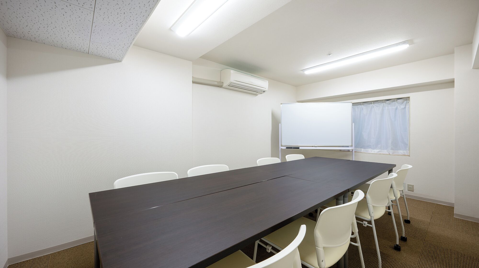Ein Konferenzraum mit langem Tisch, weißen Stühlen, einer Whiteboard und einer Klimaanlage.