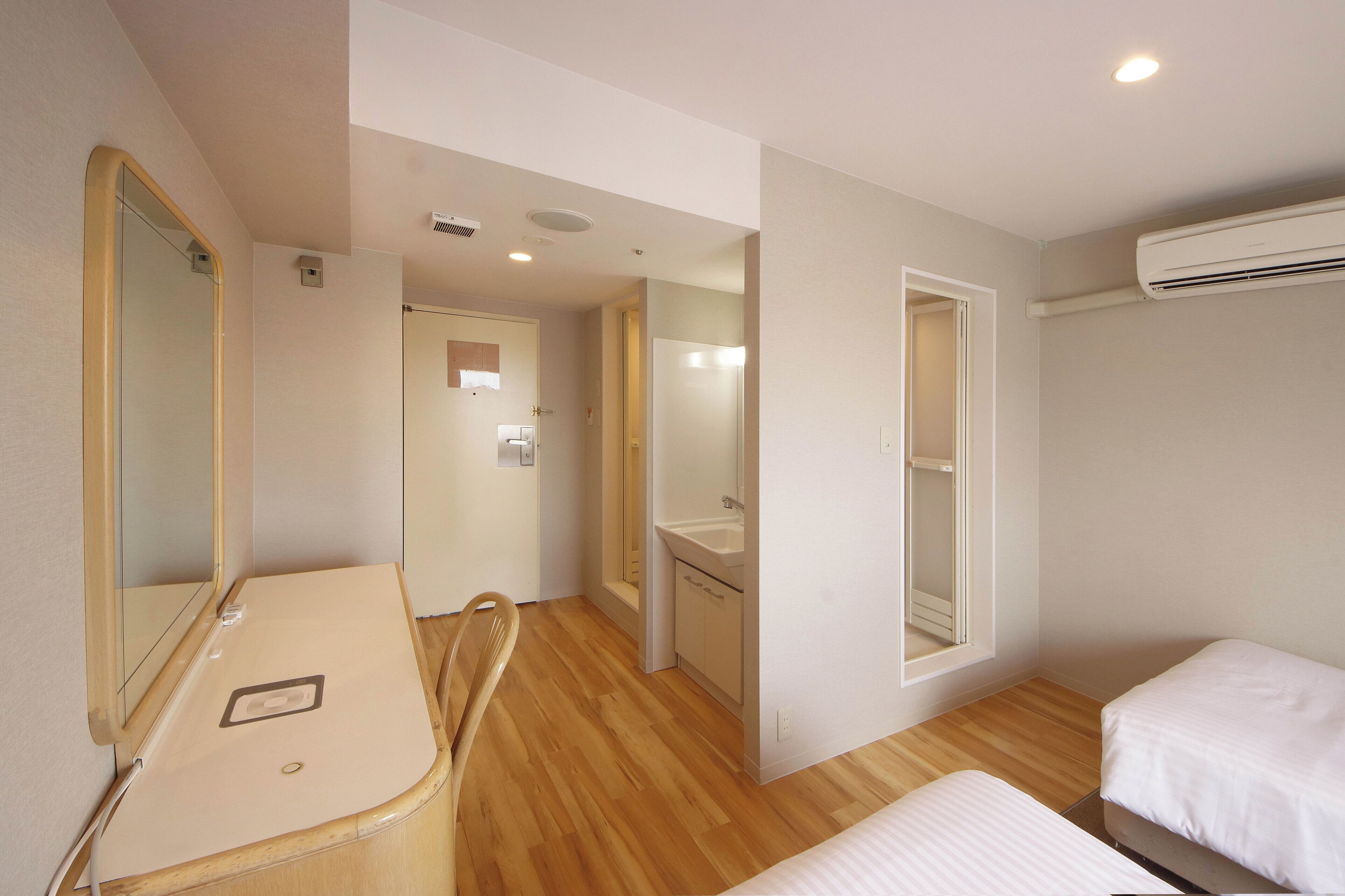 A hotel room with a wooden floor, a bed, a desk with a mirror, a bathroom with a sink, and an air conditioning unit.