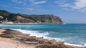 A coastal beach with rocky shoreline, clear blue water, and a hillside with buildings in the background. - Apartment- Charly in Javea (Javea)