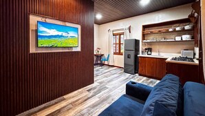 A compact living space with a sofa, a television mounted on a wooden wall, a kitchenette, and a dining area. - Tropical House (La Fortuna)