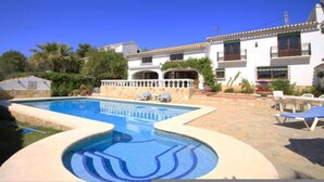 Una piscina con una fuente de agua azul cristalina, rodeada por una zona de patio con sillones de descanso y un edificio de fachada blanca y múltiples ventanales.