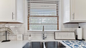 Una cocina con gabinetes blancos, un lavamanos de acero inoxidable, una ventana con persianas, un pez decorativo sobre un soporte, un dispensador de jabón y un letrero enmarcado.