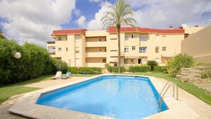 Ein Swimmingpool mit einer Palme und Liegestühlen in einem Innenhof, umgeben von einem mehrstöckigen Gebäude mit Balkonen.