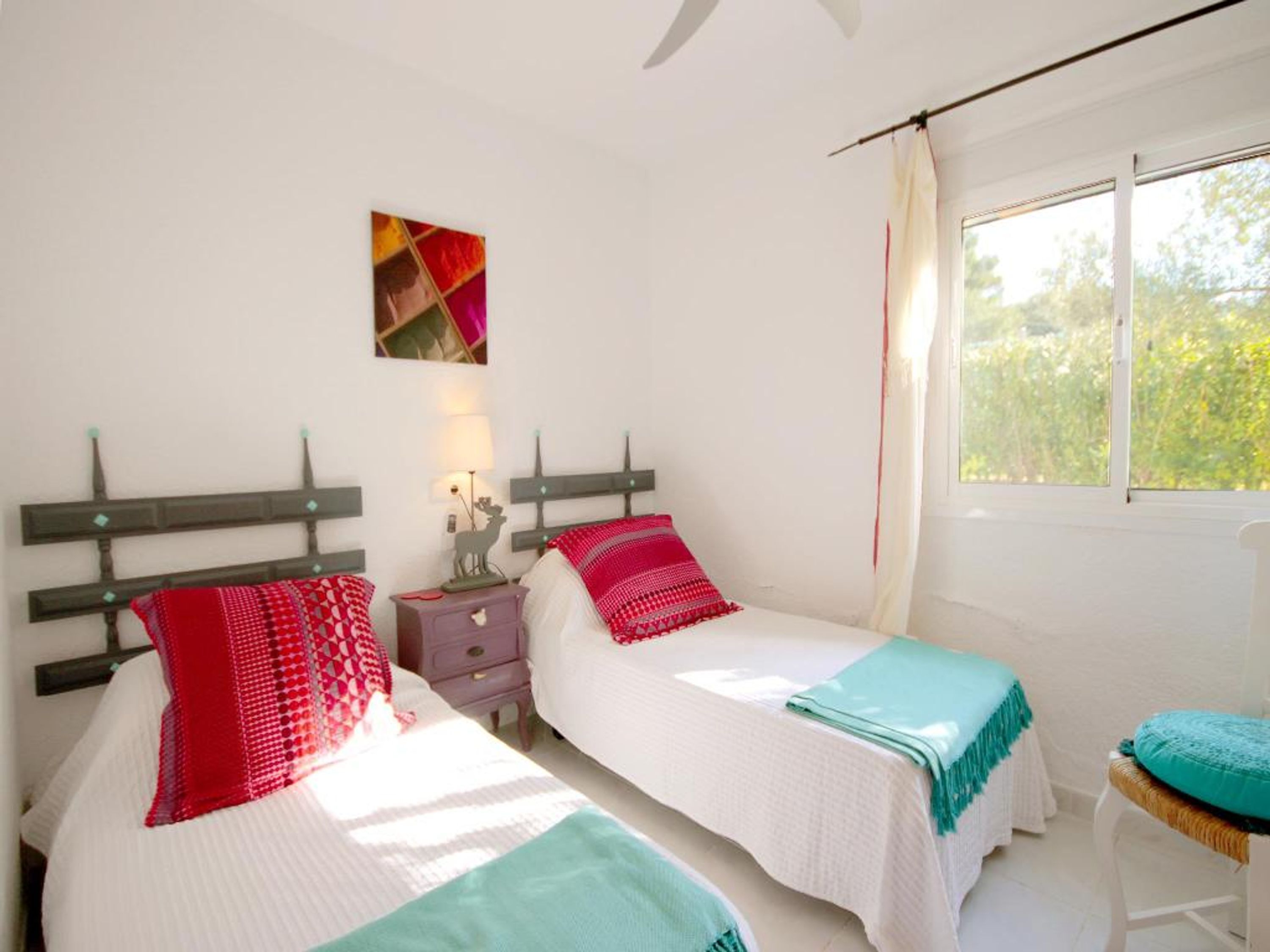 A bedroom with two beds, a nightstand, a lamp, and a window with a view of greenery.