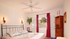 Une chambre à coucher avec un lit, un ventilateur de plafond, une fenêtre avec des rideaux, une plante et une armoire en bois.