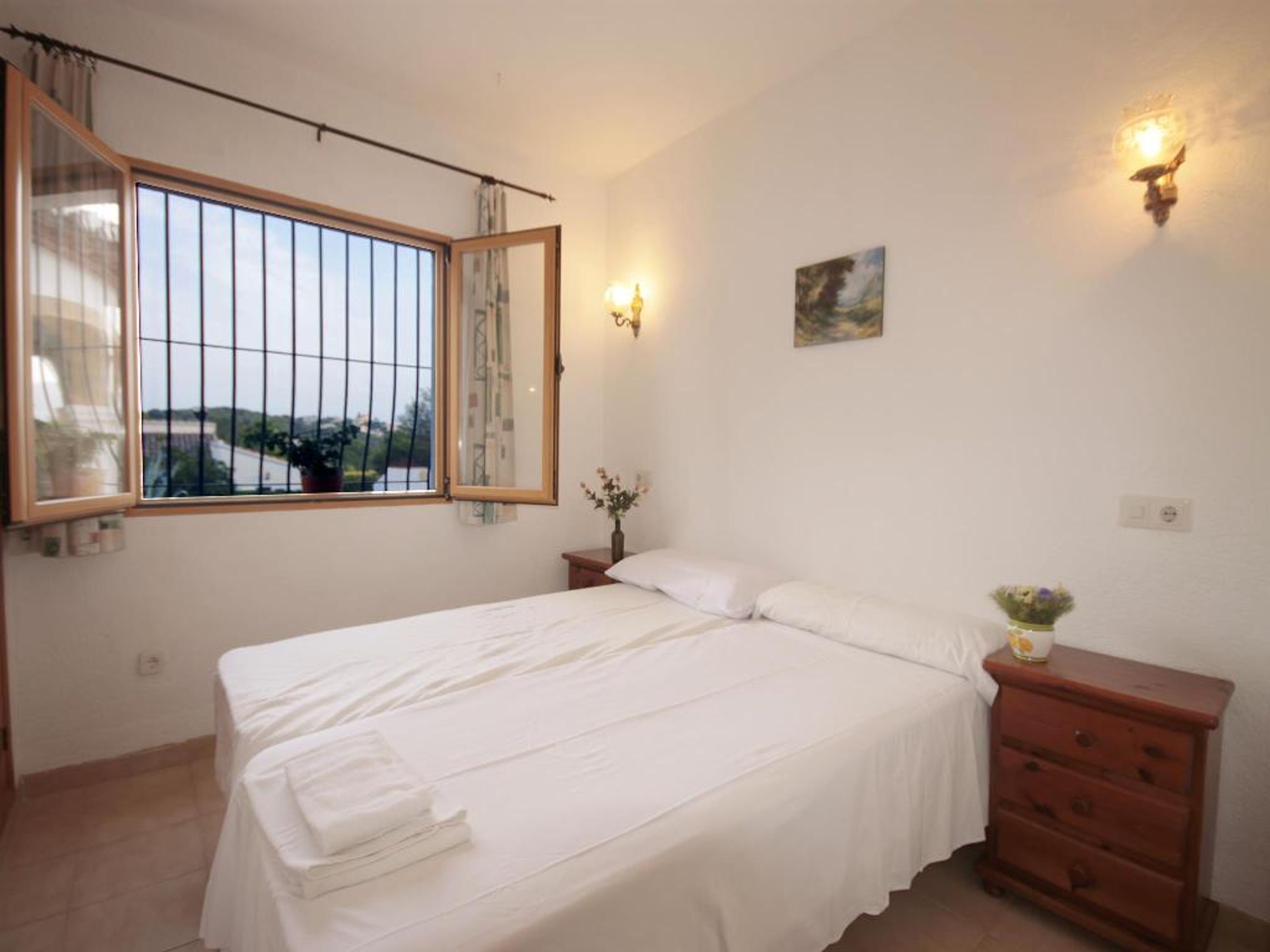 A bedroom with a large bed, a nightstand, a dresser, a window with a view, and wall-mounted lights.