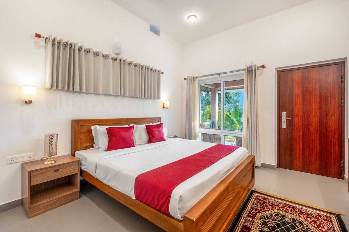 A hotel room with a wooden bed, red and white bedding, a nightstand, a window with curtains, and a wooden door.
