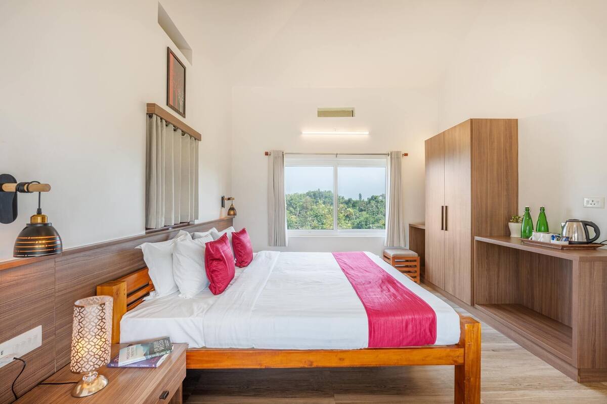 A bedroom with a large bed, wooden wardrobe, bedside table with lamp, and a view of greenery through the window.