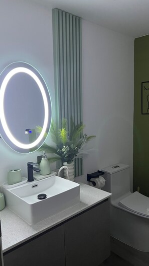 Un baño con lavamanos blanco, un espejo con luz, un inodoro y una pared verde.