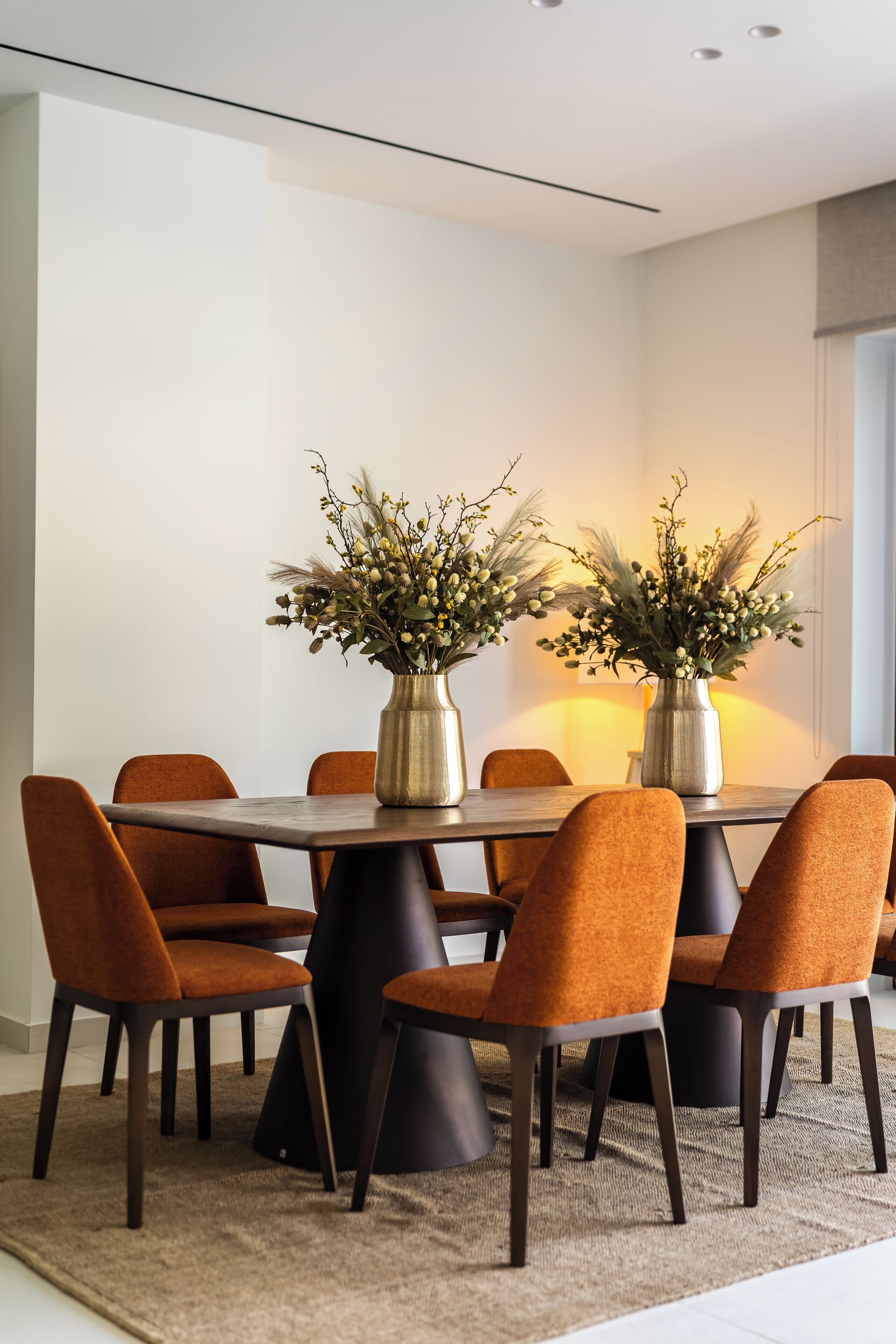 A dining area with a round table, six chairs, two vases with flowers, and a light source on the wall.
