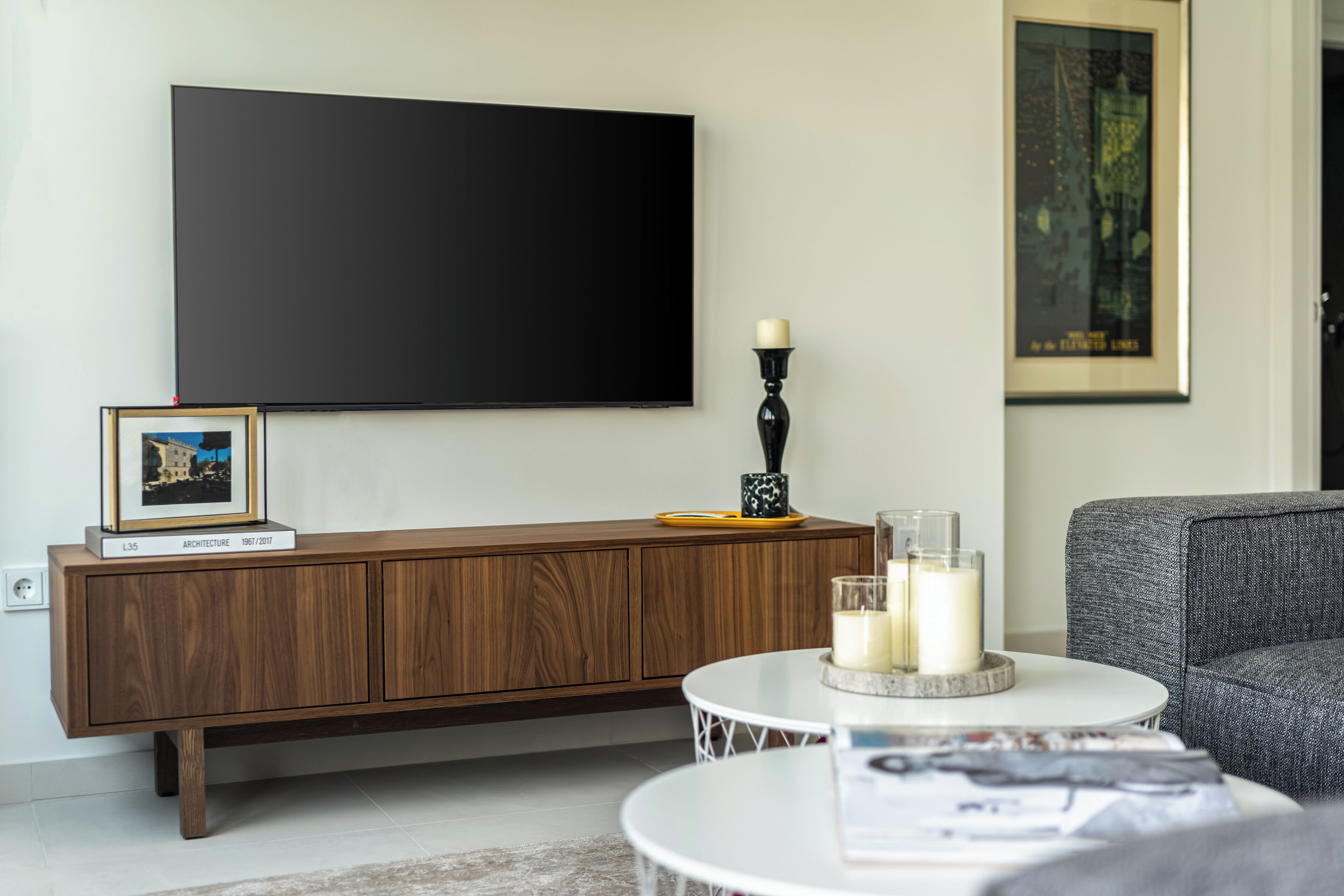 Een moderne woonkamer met een grote flatscreen televisie aan de muur, een houten kast, een ronde salontafel met kaarsen en een lijst met een foto op de kast.