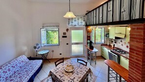 A compact kitchen with a gas stove, a dining table with chairs, a small window with a view of greenery, and a bed with a floral bedspread. - Casa Ariel in the historic village. (Rio dell'Elba)