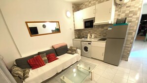 Un espace de vie compact comprenant un canapé, une table basse en verre, un lave-linge et un coin cuisine de taille réduite.