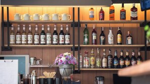 Een bar met flessen drank, koffiemokken en een bloemstuk.