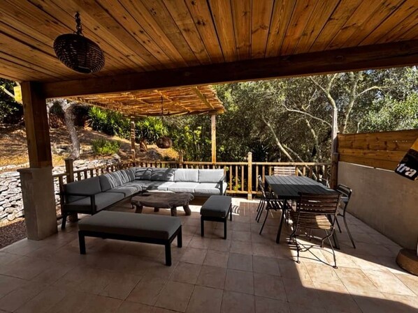 An outdoor patio with a wooden pergola, a sectional sofa, a dining table with chairs, and a stone wall.