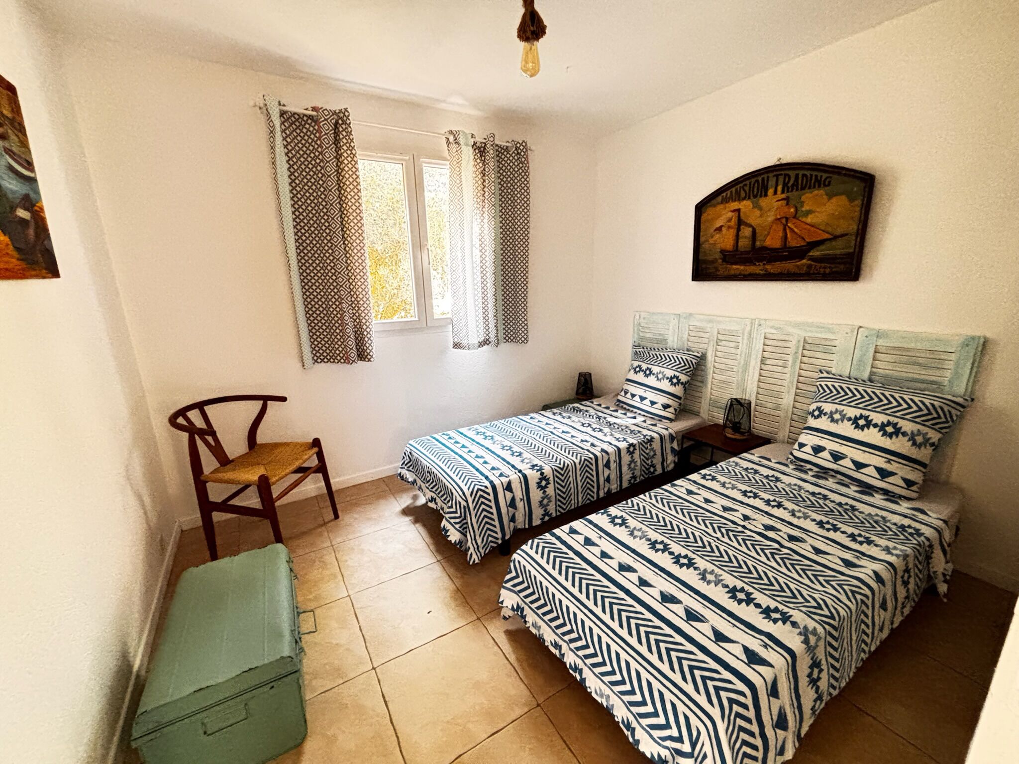 A small, clean room with two single beds, a chair, a small table, and a window with patterned curtains.