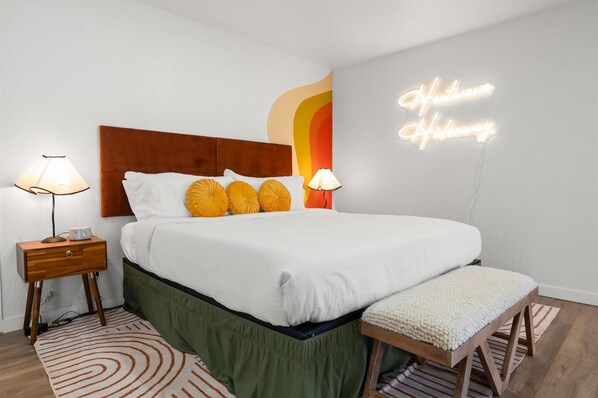 Un dormitorio ordenado con una cama grande, dos lámparas de noche, una mesita de madera, un banco y un letrero de neón en la pared.