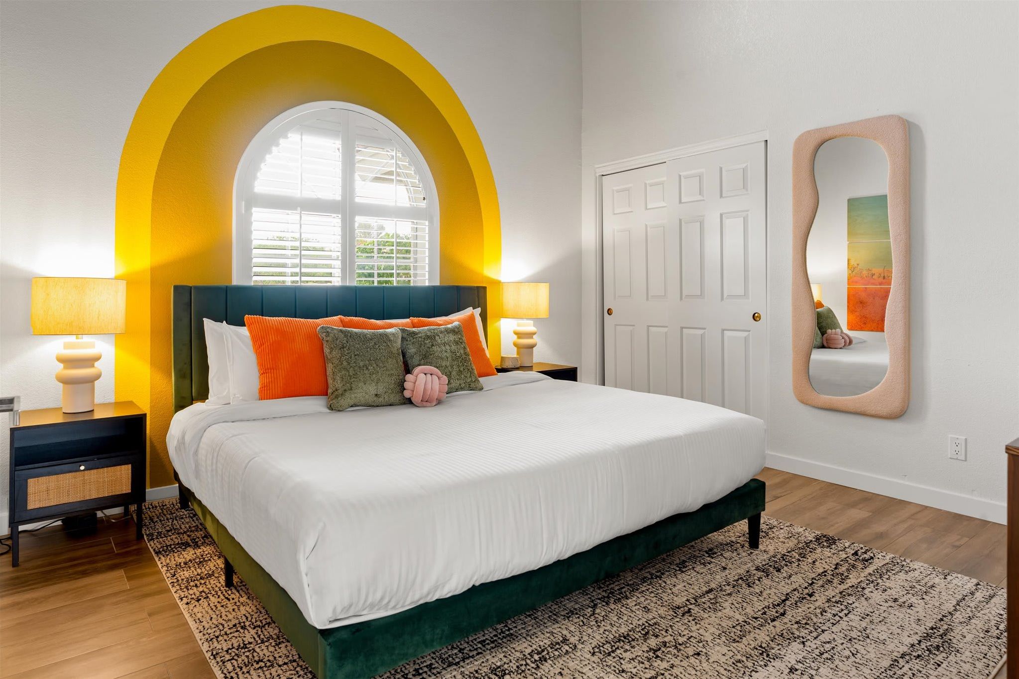 A bedroom with a bed, two bedside tables, a mirror, and a window with a yellow arch.
