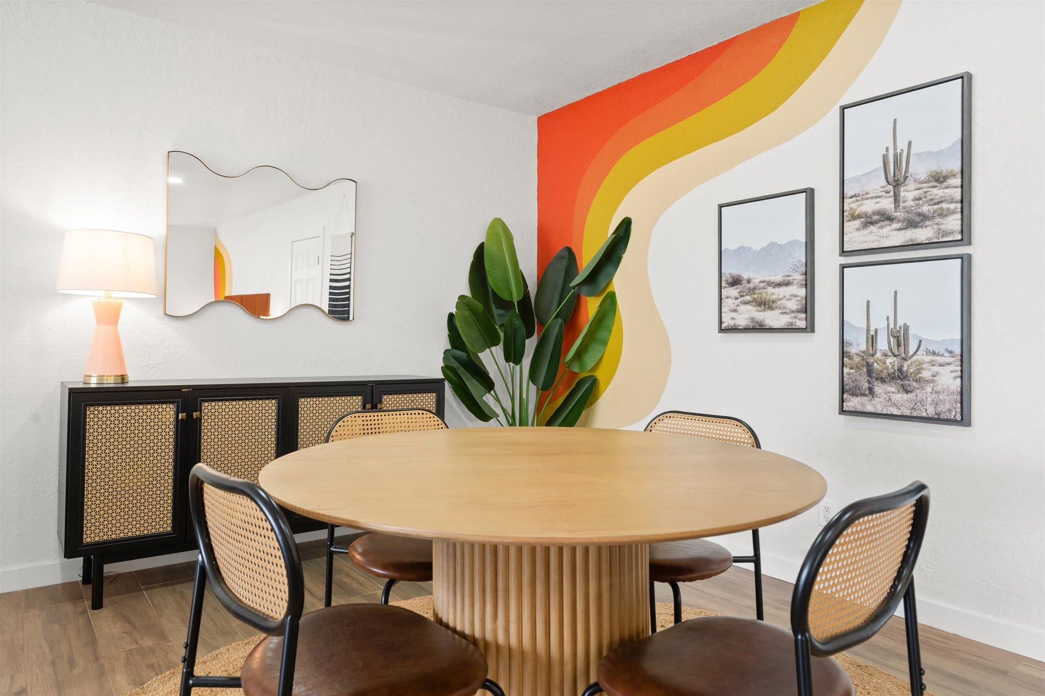 A dining area with a round wooden table, black chairs, a woven rattan bench, a modern lamp, and a large abstract painting.