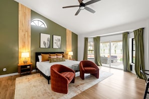A bedroom with a bed, two armchairs, a ceiling fan, a nightstand with a lamp, and a large window with curtains.