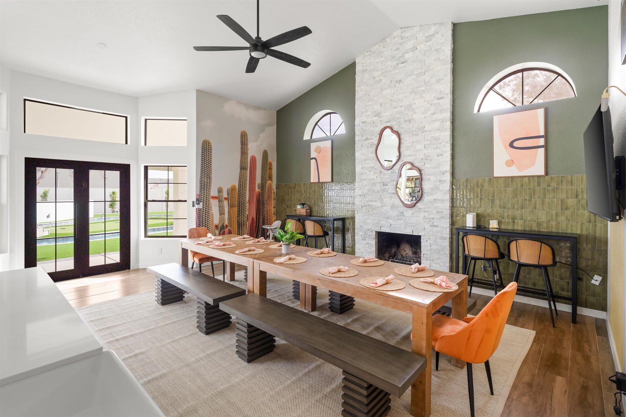 Un espace de salle à manger moderne avec une longue table, des bancs et des chaises. On y trouve une cheminée, un ventilateur de plafond et une grande fenêtre donnant sur un paysage verdoyant.