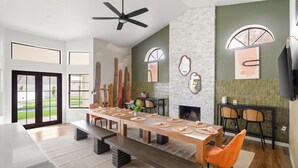 Un comedor moderno con una mesa larga, bancos y sillas. Hay una chimenea, un ventilador de techo y un ventanal con vistas a un paisaje verde.