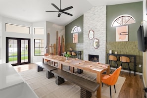 A modern dining area with a long table, benches, and chairs. There is a fireplace, a ceiling fan, and a large window with a view of a green landscape.